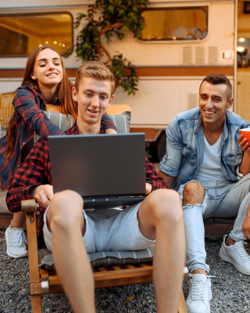 A man and woman smiling while watching and using the laptop with RV parked at the background.